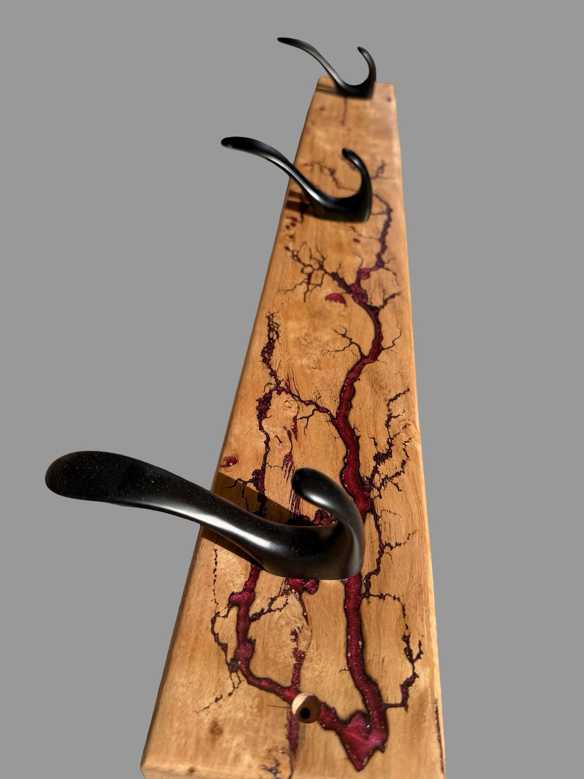 A rustic wooden coat rack with a natural wood grain base featuring dark red and black crackle inlay patterns, holding three black metal hooks against a solid gray background.