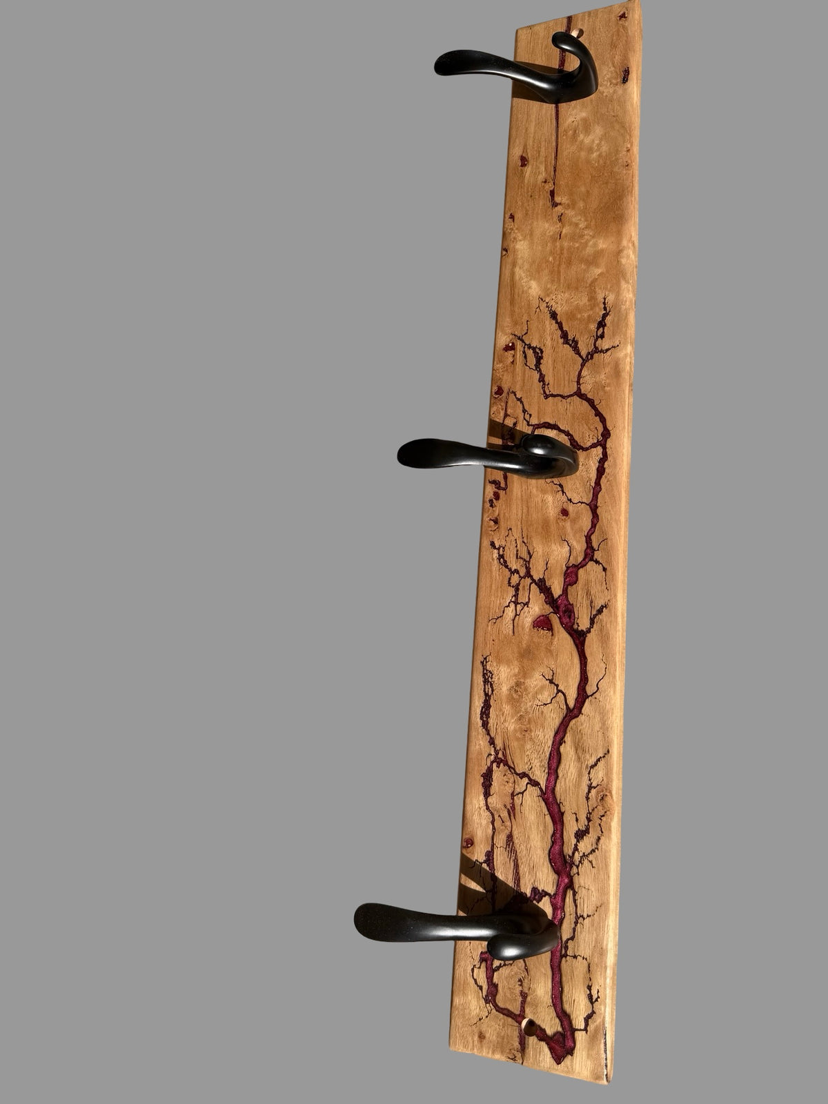 A rustic three-hook coat rack made from a natural wood panel with dark brown grain patterns and three black metal hooks, shown against a solid gray background.