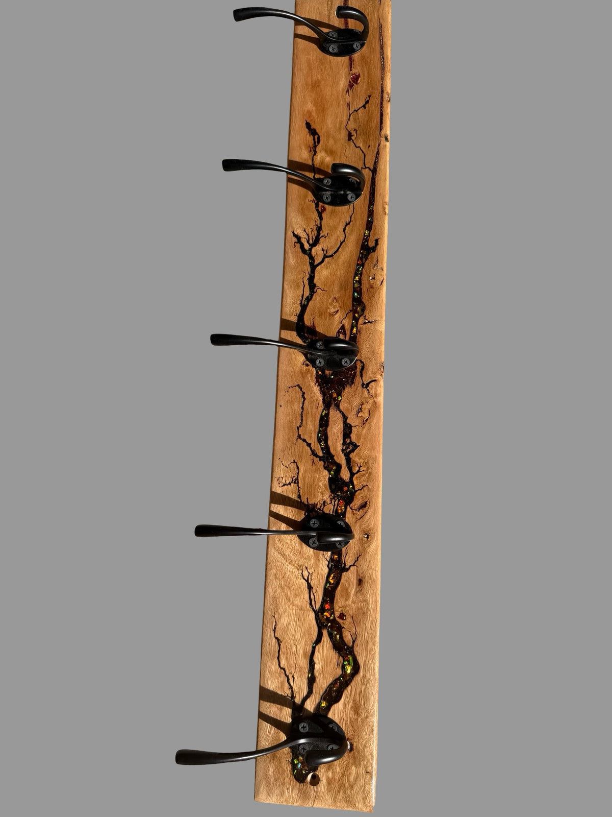 A rustic wooden coat rack with five black metal hooks, featuring a natural live-edge wood grain pattern with dark burn marks, shown against a solid gray background.