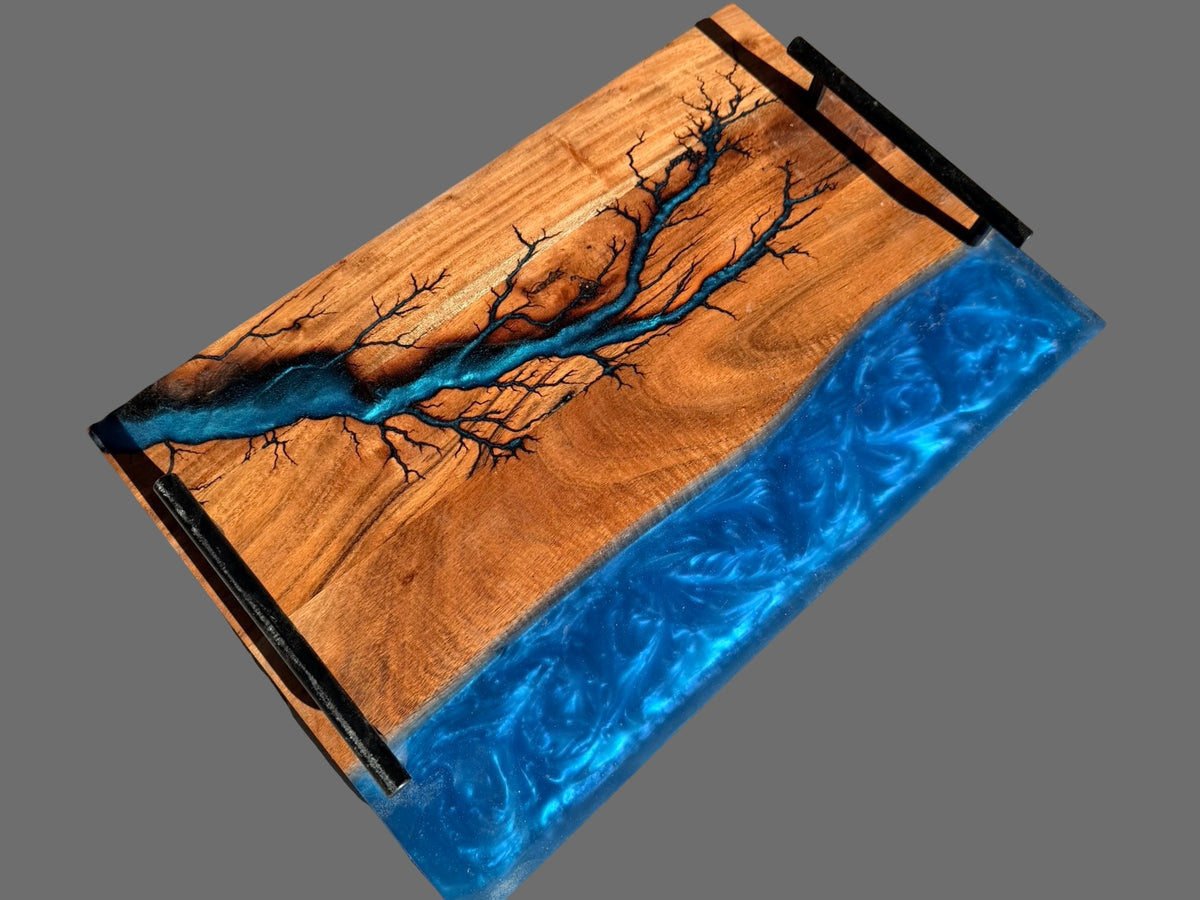 A rectangular wooden resin serving tray with black metal handles, featuring a natural wood grain top with a branch-like pattern and a bright blue resin river design along one side, shown on a solid gray background.